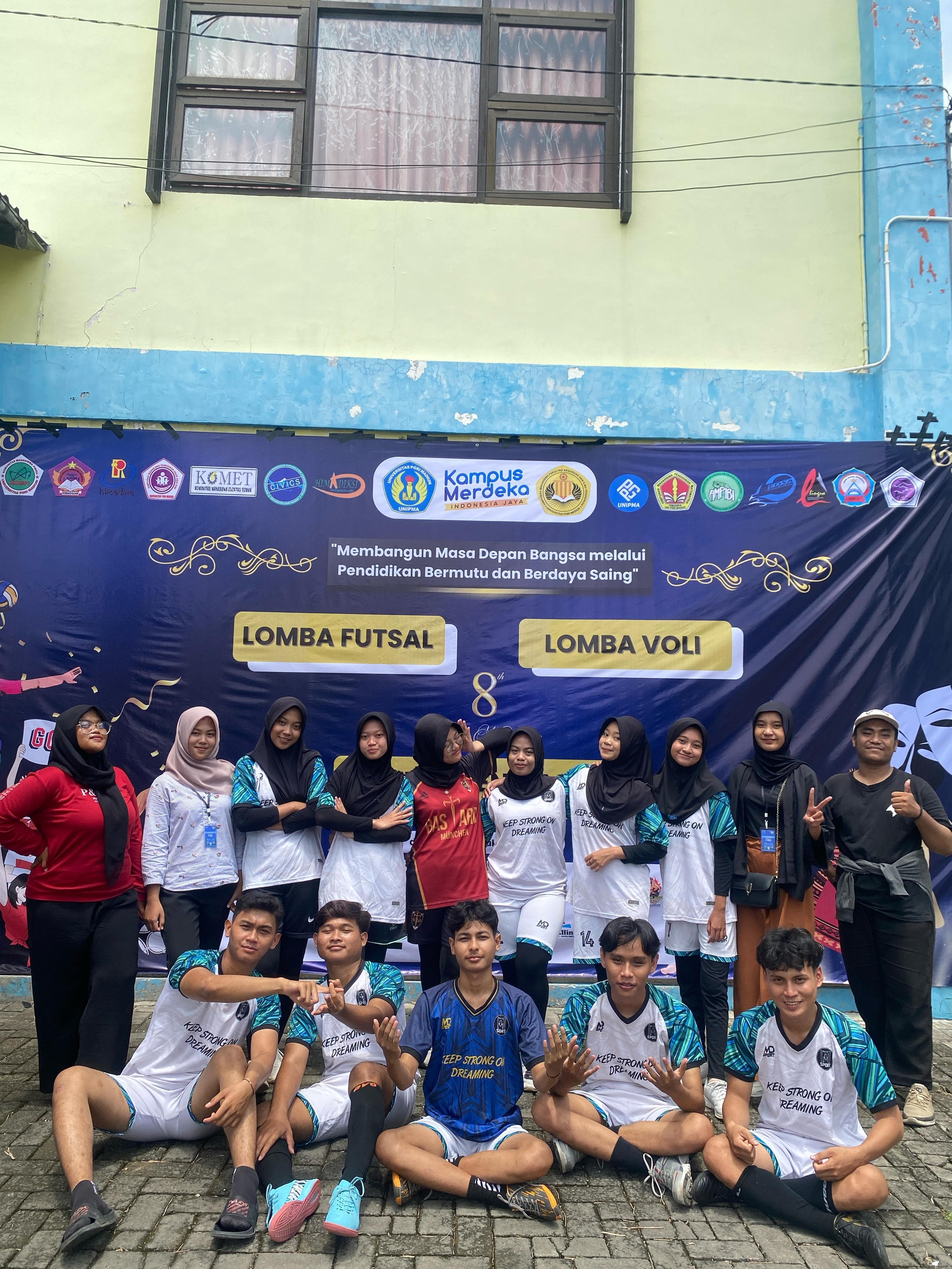Young students, some in sports jerseys, pose smiling before an Indonesian university event banner.