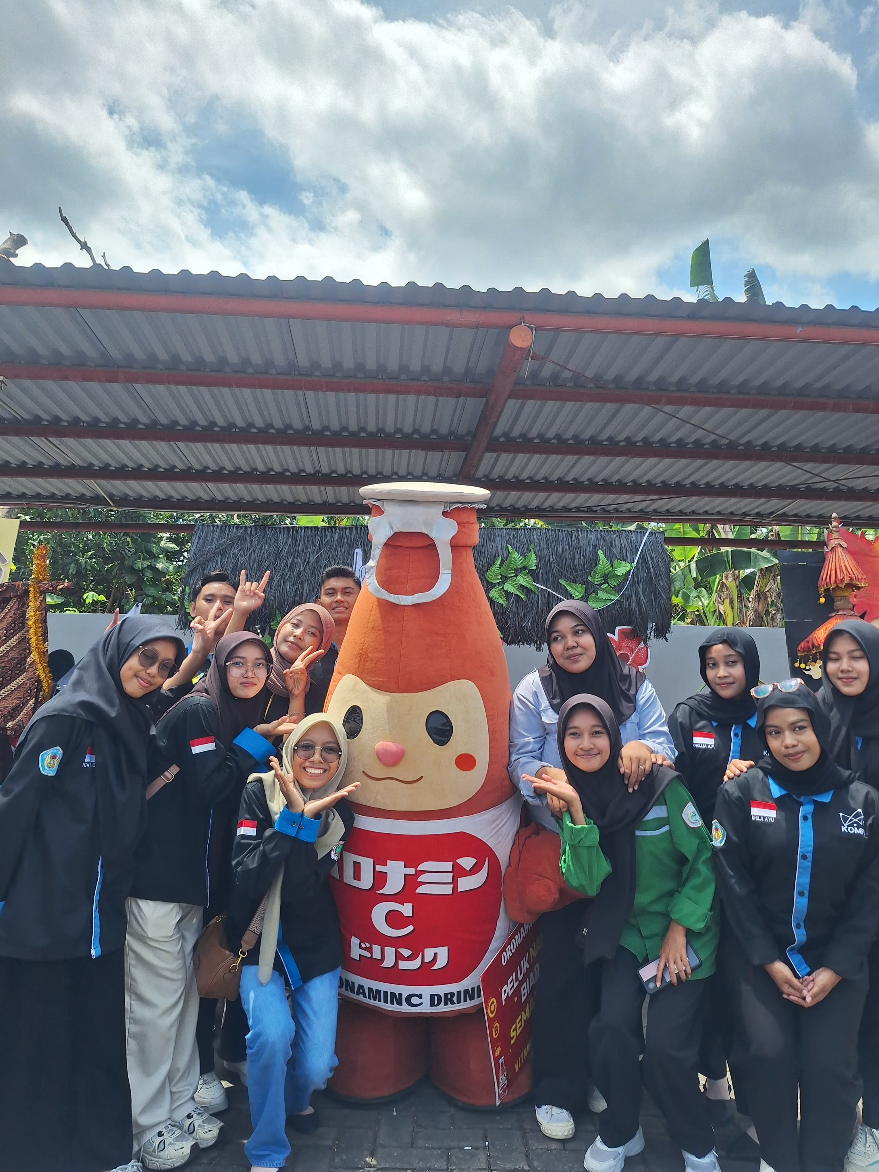 Group of smiling young people posing with a giant Oronamin C drink mascot outdoors.