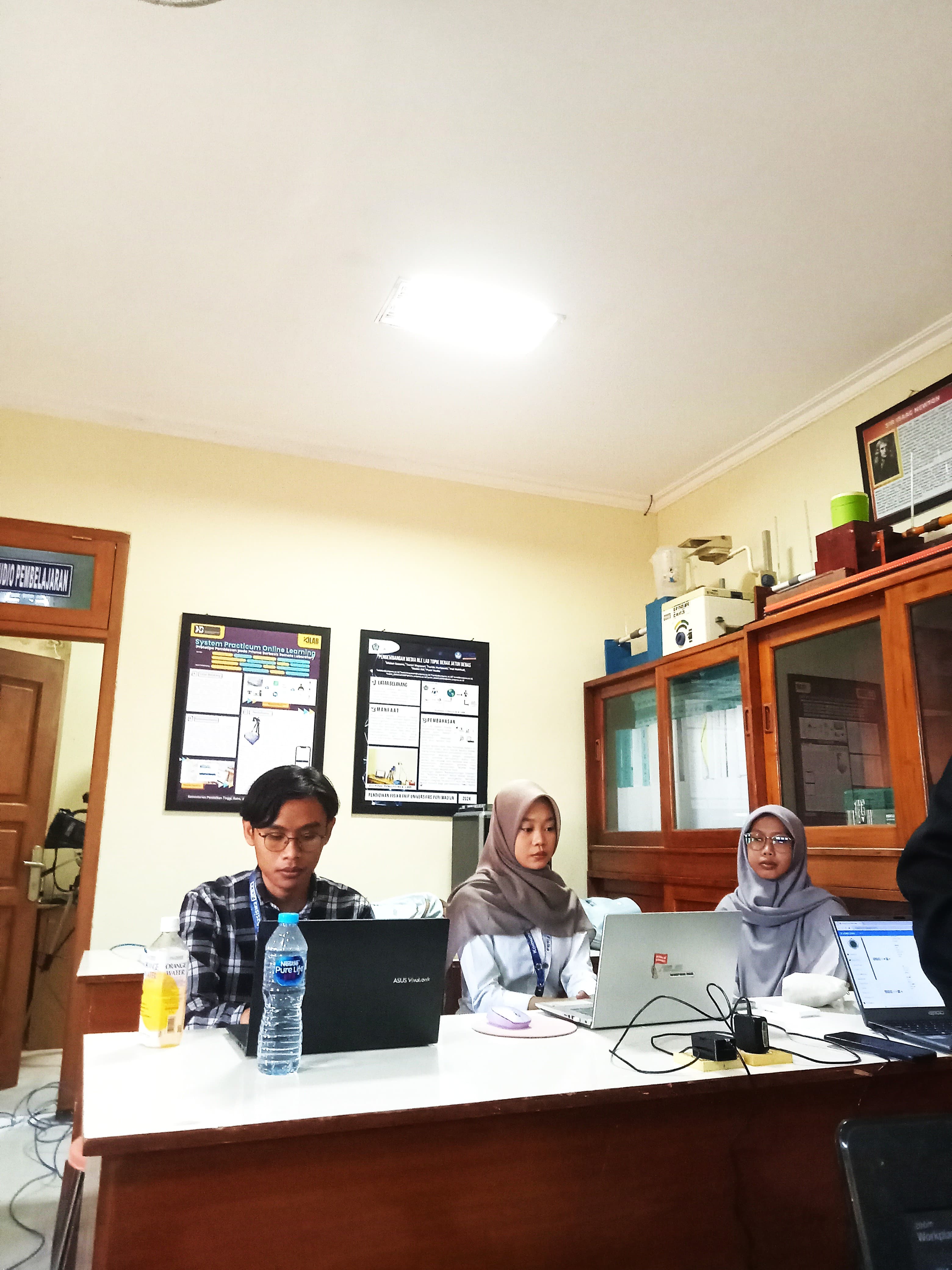 Students, two wearing hijabs, working on laptops in a room with scientific posters.