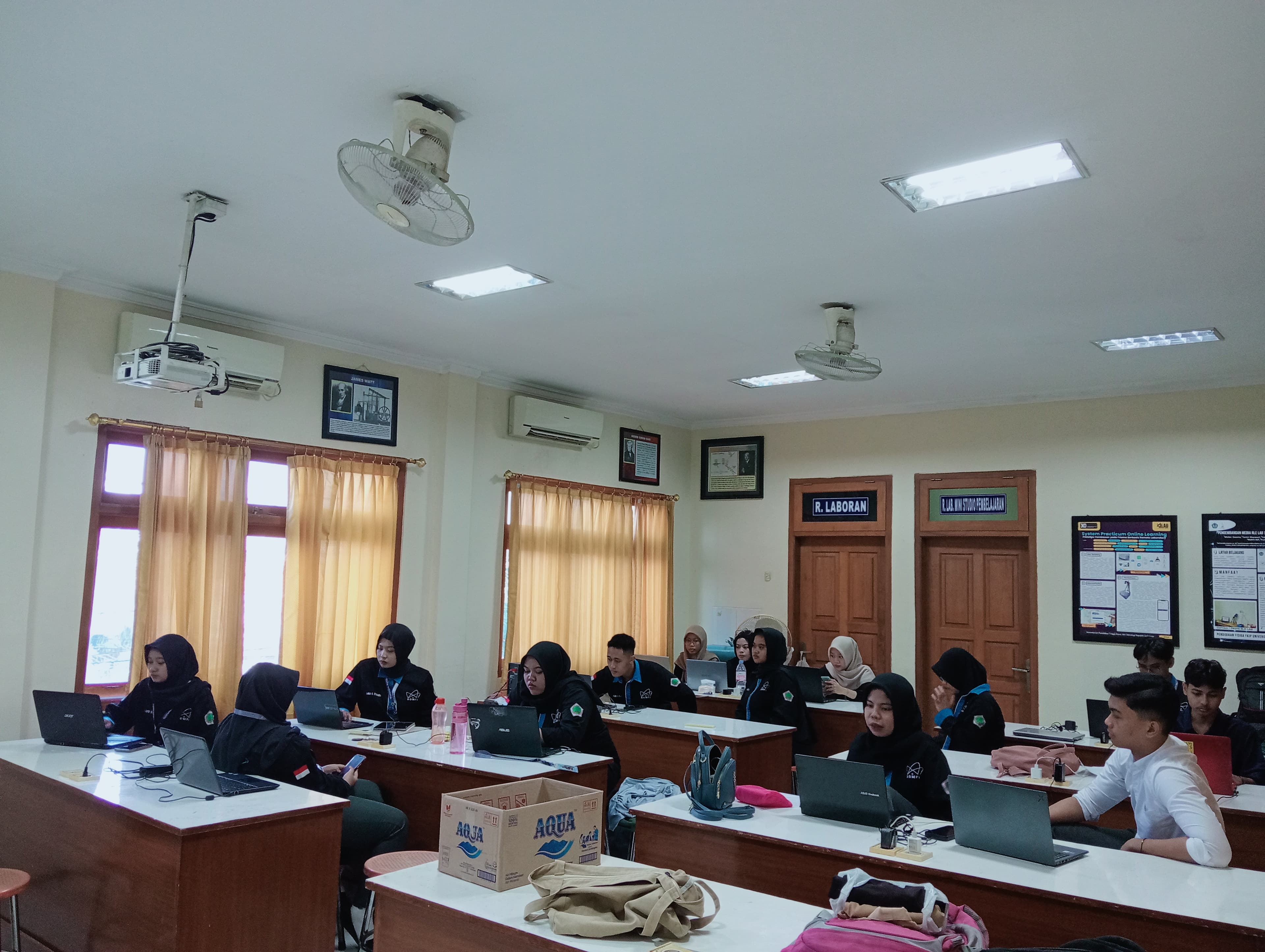 Young adults in a bright classroom, focused on laptops at individual desks.