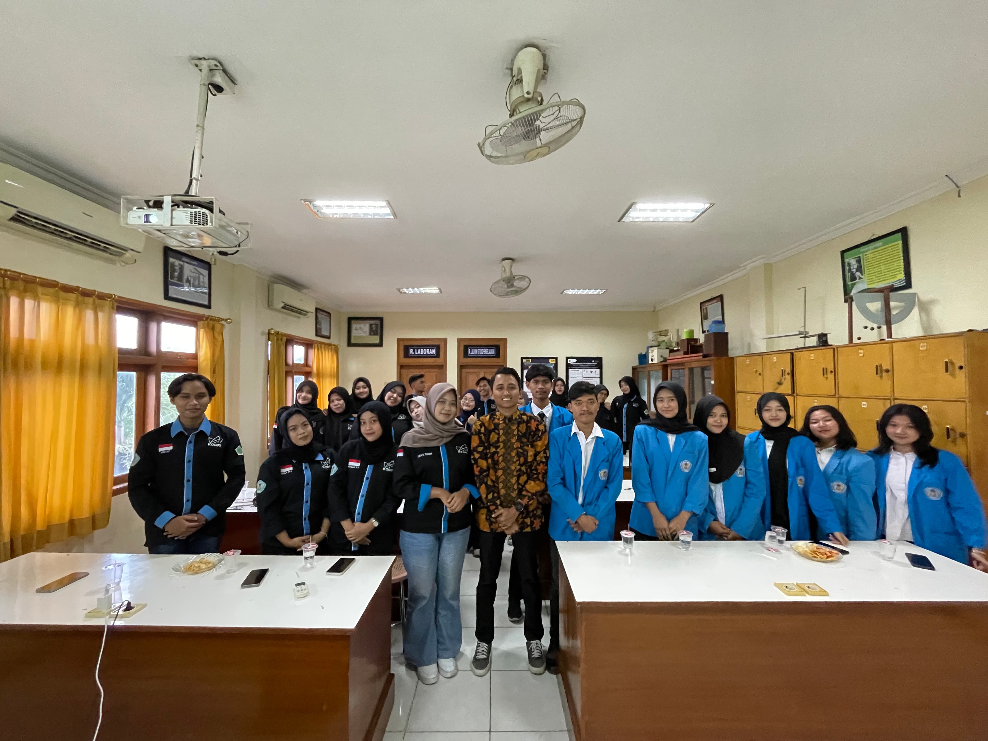Young adults in blue jackets and batik shirt standing in a bright classroom.