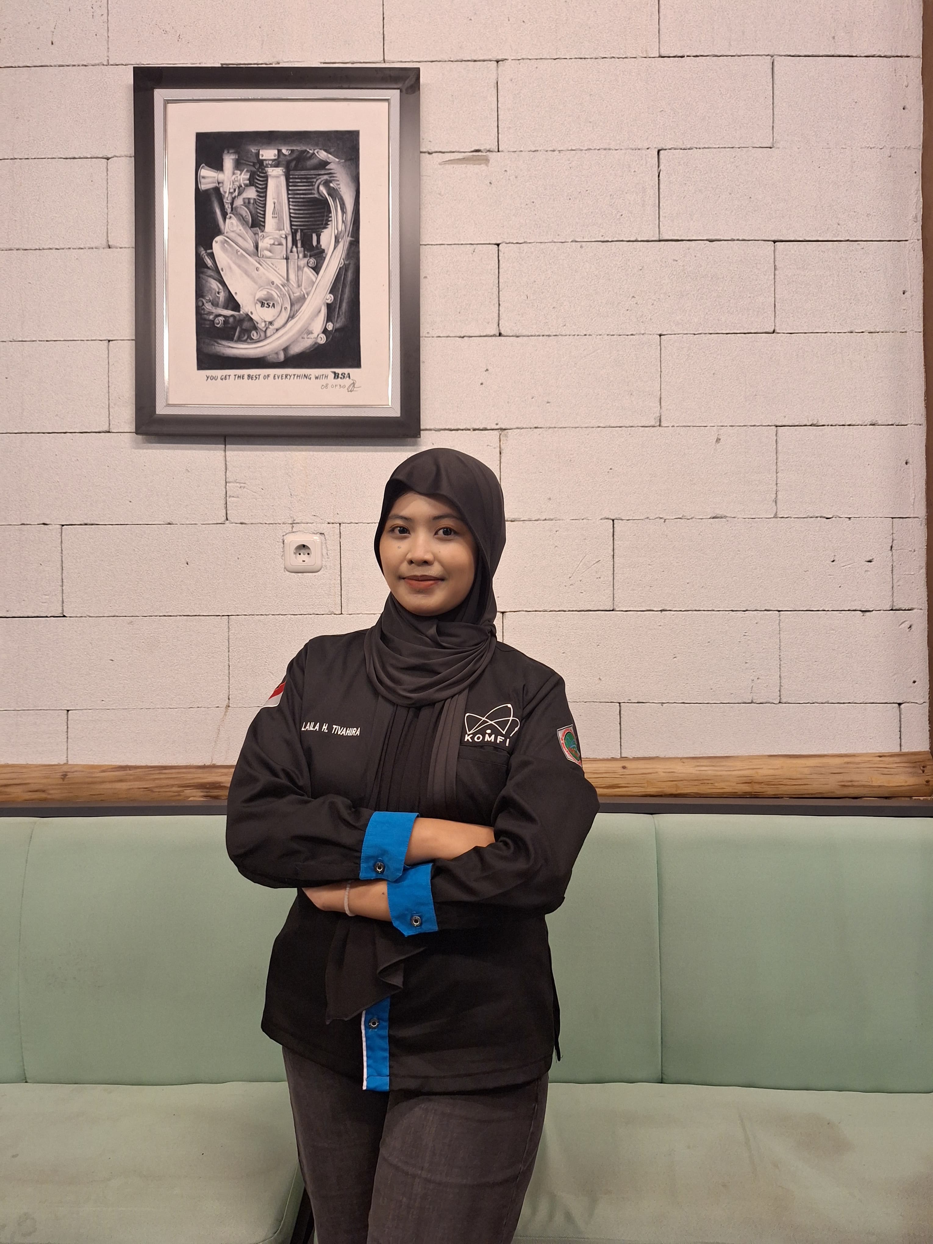 Smiling woman in black hijab and jacket, arms crossed, against a white brick wall.