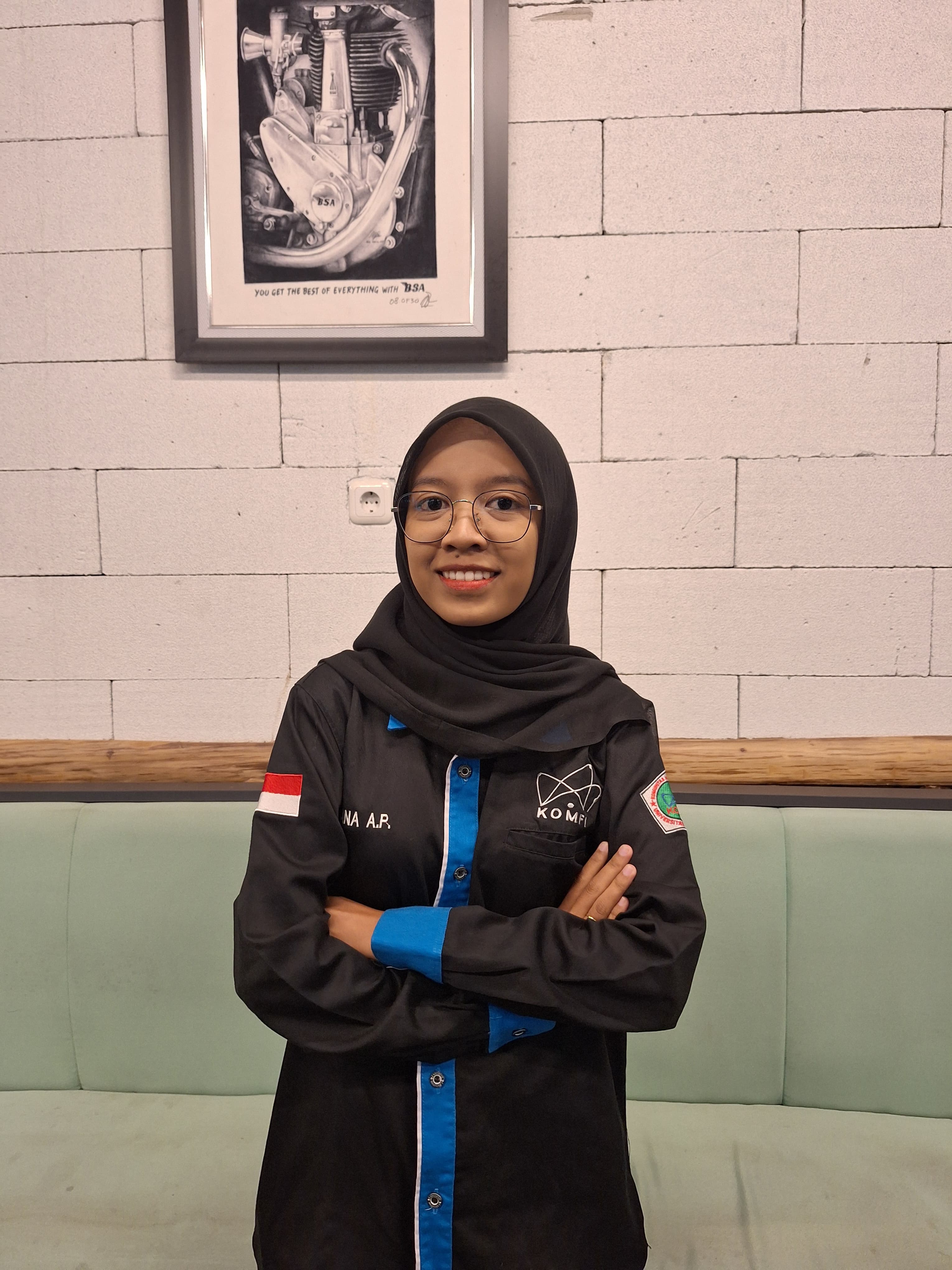 Smiling woman in black hijab, glasses, and blue-accented jacket, arms crossed, beside motorcycle art.